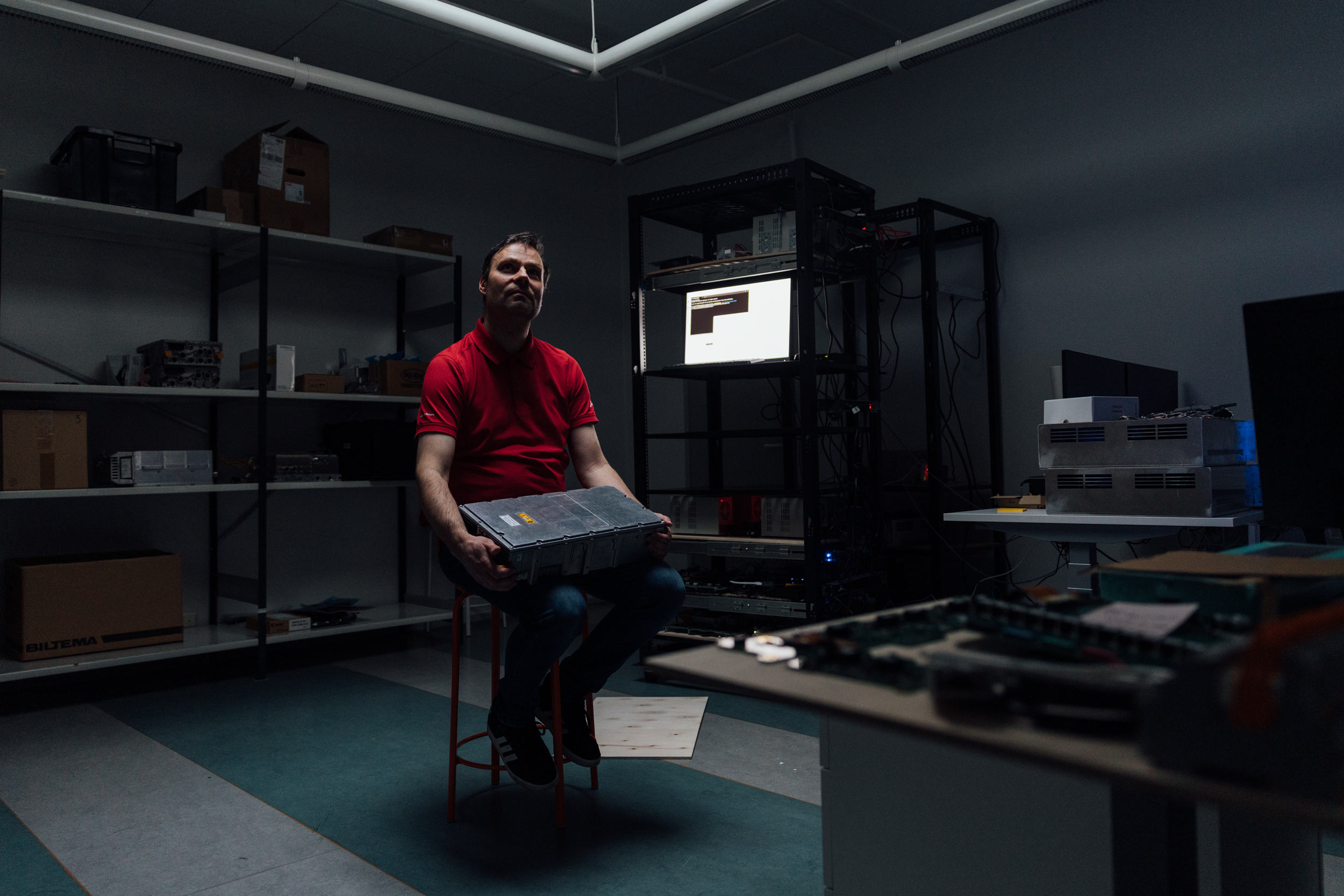 Mies punaisessa t-paidassa istuu hämärässä tehdastilassa pitäen käsissään laatikkomaista laitetta. Taustalla teollisuustilan hyllyjä, näyttö, työpöytä.